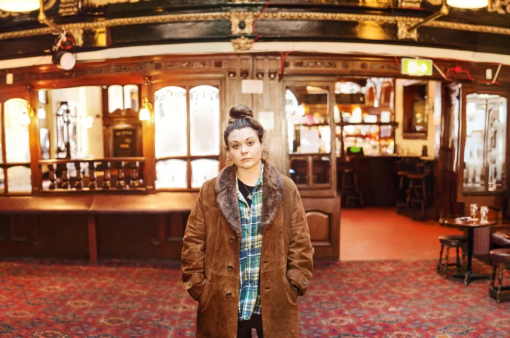 Maria Ferguson stands inside an old pub wearing a blue checked shirt and long brown coat.