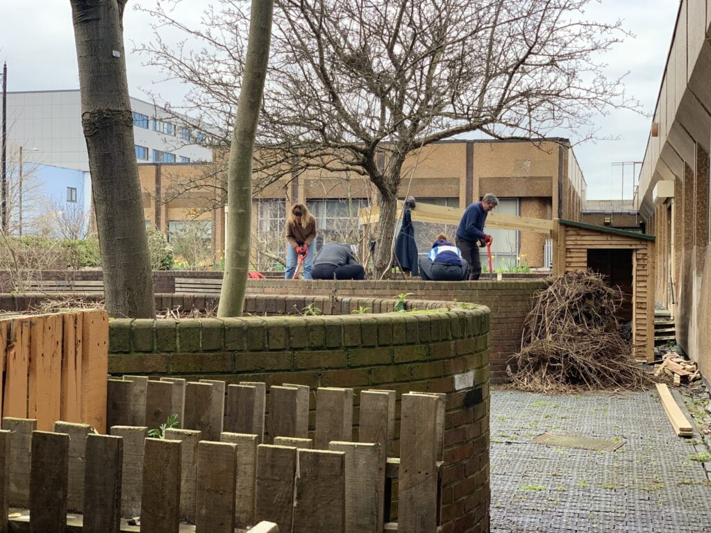 People gardening outside a small shed, in an area enclosed by brick walls. A wooden fence in the foreground, and leafless, winter tree in the background.