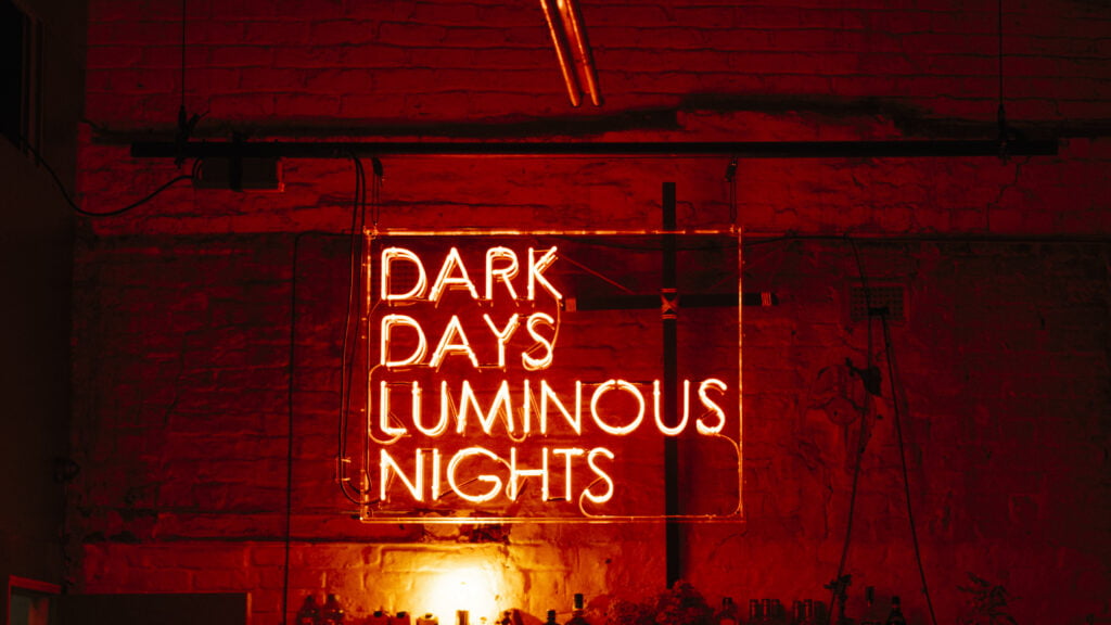 A red neon sign with the words 'DARK DAYS LUMINOUS NIGHTS' against a brick wall.
