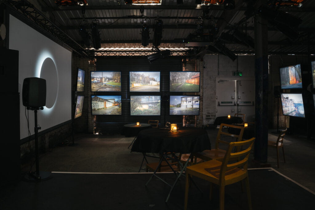 Dark room with a large screen on the left and smaller lightboxes in the centre, where photographs of Manchester are displayed. There are tables in front of it, with black tablecloth, candles and chairs around it. 