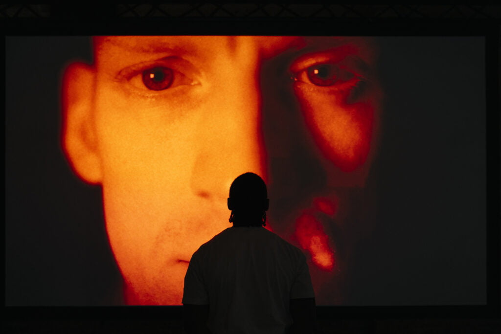 Face of a man on a large screen, lit up by red light. There is a silhouette of a man in front of it, with his back to the camera.