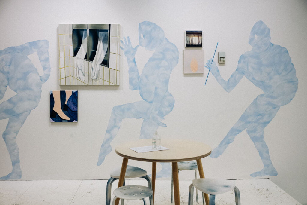 Pale blue paintings of strange human-like creatures on a wall, next to rectangular canvases depcting a window, feet and a curtain. In front of the wall there is a round wooden table and stools.