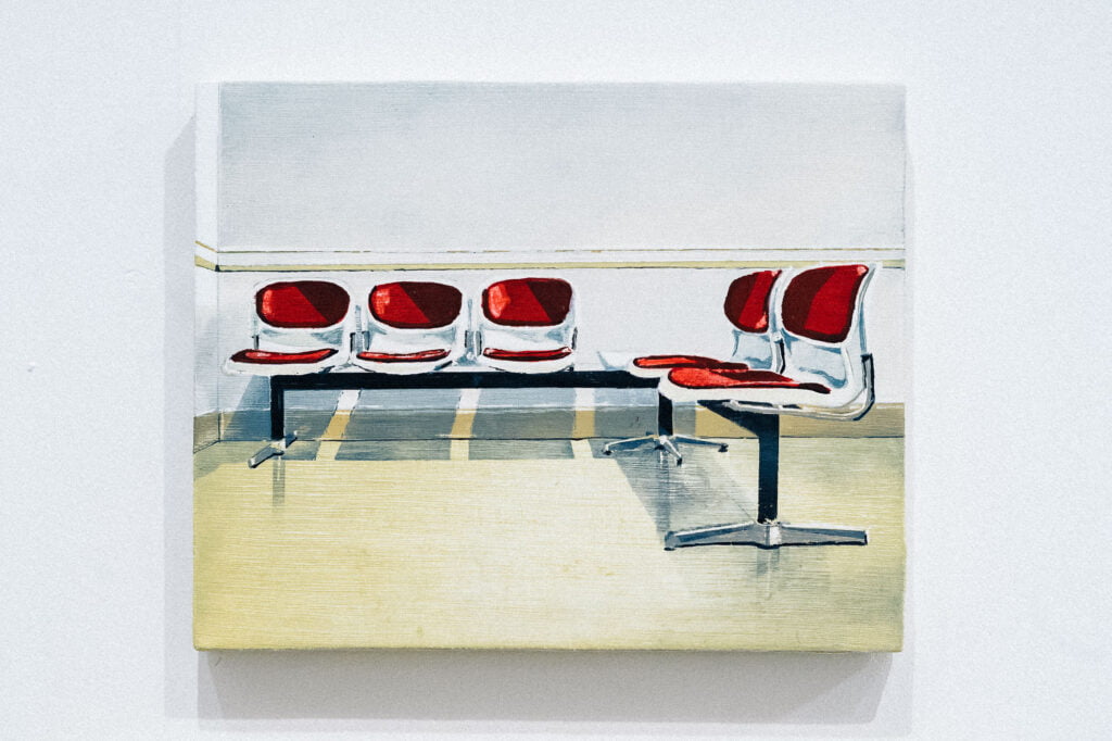 A painting by Richard Baker of a row of seats, similar to what you would find in a waiting room, side by side, joined by a metal frame, with red seats and backrests on each seat, in white casing. The floor is pale yellow and the wall behind them is white.