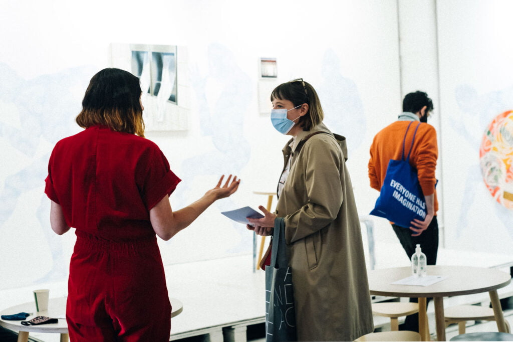 Two women talking in the exhibition space, one is wearing a red boiler suit and has shoulder length dark hair wtih blonde ends and the other is wearing a sand-coloured coat with a hood and a medical facemask. There is a man looking at the art in the background, wearing an orange jumper and holding a blue tote bag with the words 'Everyone has imagination'.