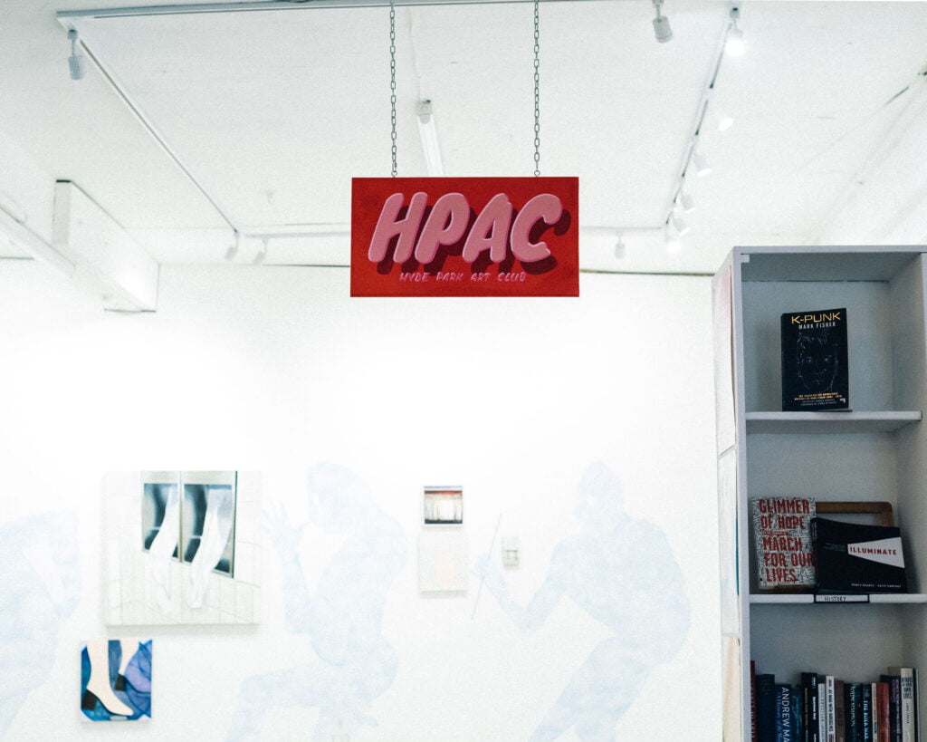 Sign of the Hyde Park Book Club in pink letters on red background, suspended from the ceiling of a white room