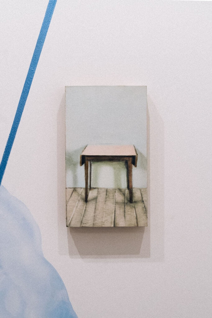 Painting of a table against a white wall, standing on a wooden floor