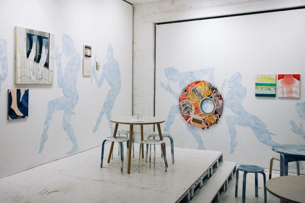 Cornern of a room with blue figures on the wall, surrounded by painted canvases, with a stage and white tables and stools in front.