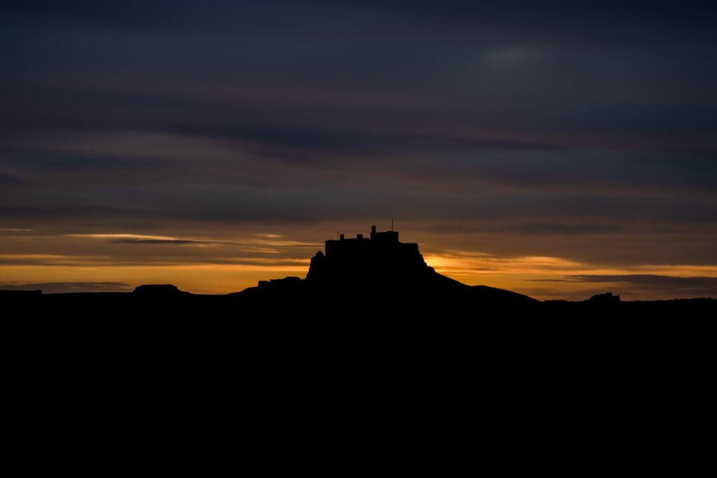 Silhouette of a castle on a hill against a sky during sunrise, going from a strip of bright yellow into a cloudy blue-grey.