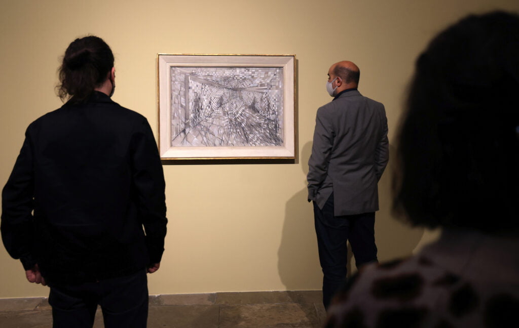 3 people seen from the back looking at a painting of an abstract black and white composition in a white and gold frame on a pale yellow wall.