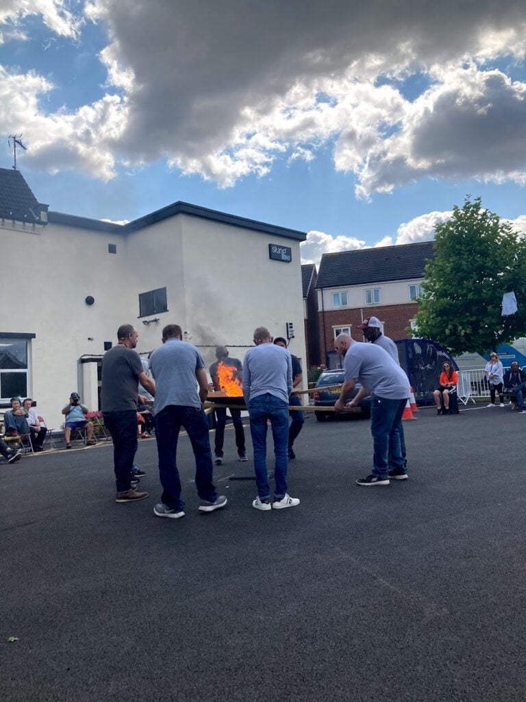 A group of men, dressed in blue trousers and long sleeved tops around a fire, ourtside a white building on the pavement, below a cloudy sky