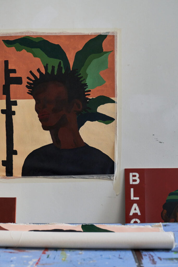 Studio view of a painting in progress. The canvas is on the wall and depicts a black man with spiky hair and a leafy pattern in the background.
