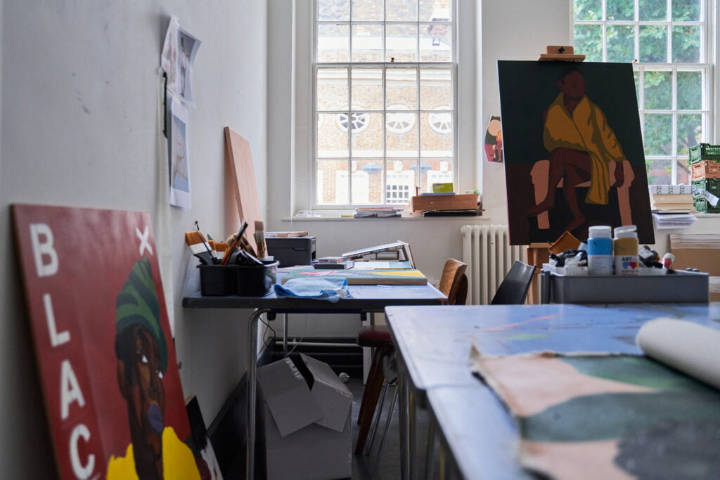 Artists studio with one painting on an easel, and another one propped against the wall. There are big windows and nautral light coming in, there is a table at the front with a painting in progress.