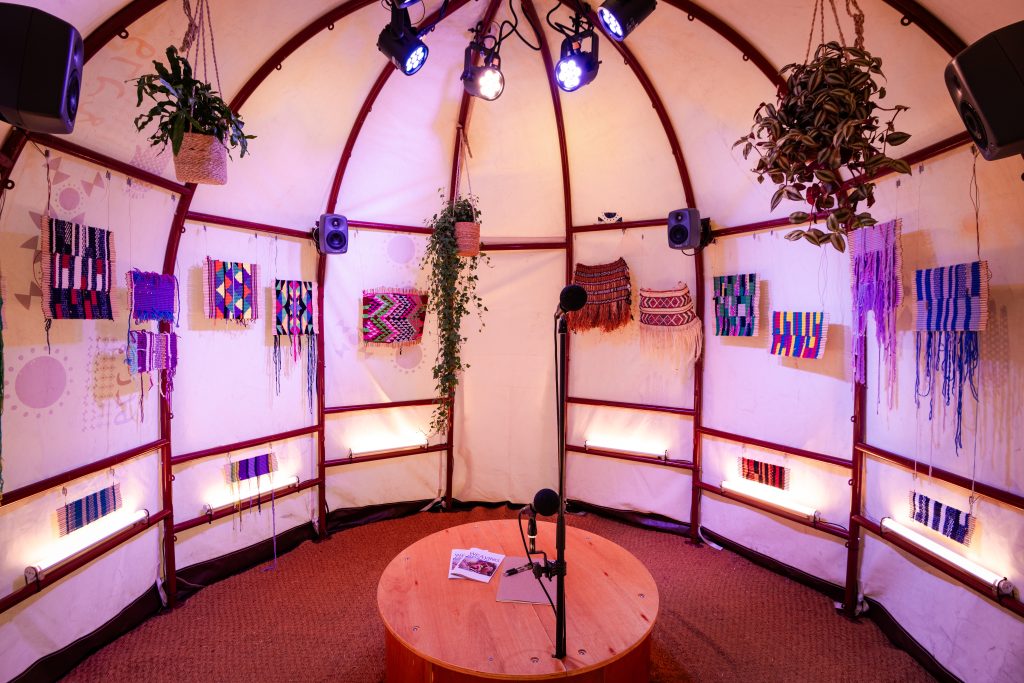 Image of the interior of the aqal, with tapestries on the walls, plants hanging from the ceiling and a stage with a microphone in the centre.