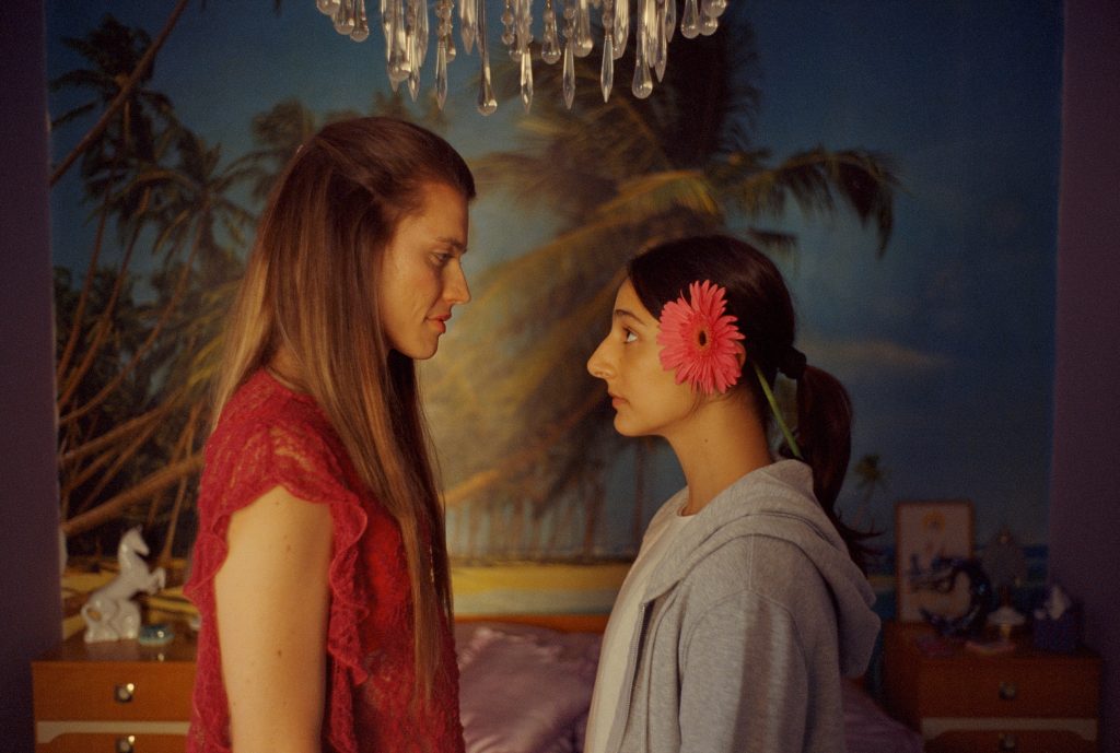 Two young women stand close, facing each other in a bedroom with a tropical wall mural; one in a red dress, the other with a pink flower in her hair.