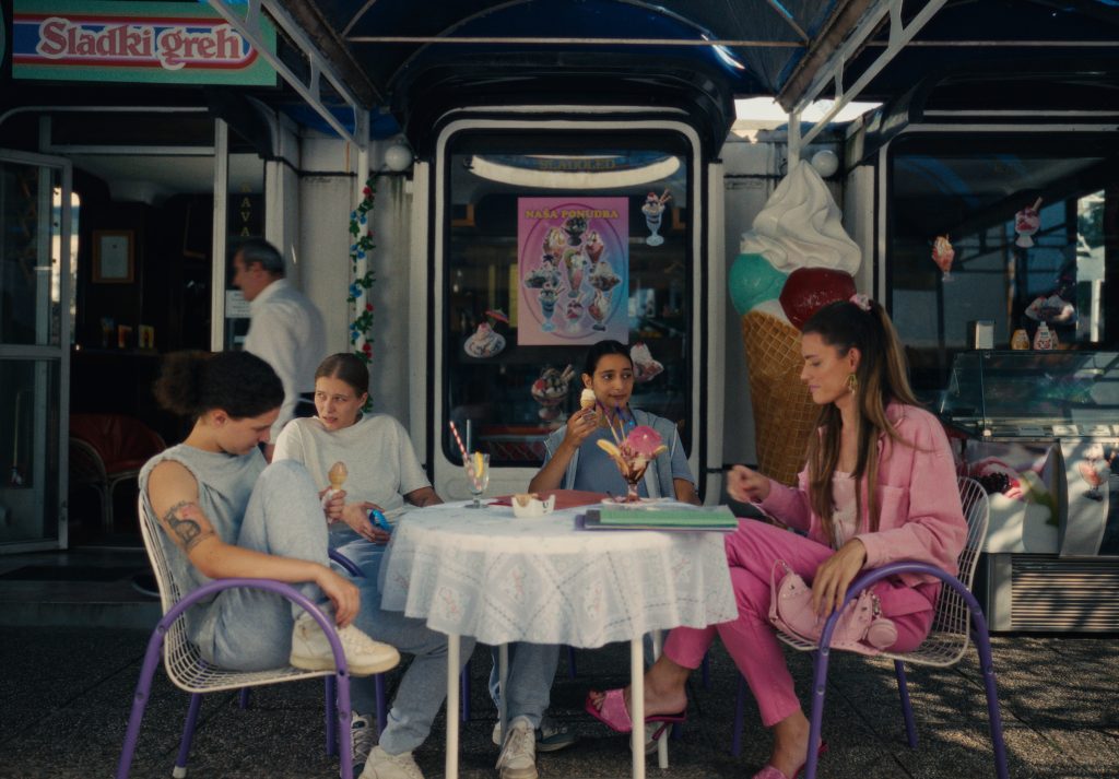 Four young women eat ice cream at an outdoor café, seated around a small table in front of a brightly decorated shop with a giant ice-cream-cone display.