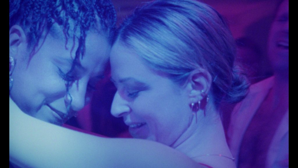 Two women dance closely in blue and purple club lighting, their foreheads touching and eyes closed.