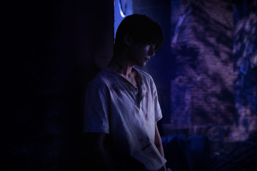 A person in a dim, bluish-purple lit scene leans against a wall, looking downward with a somber expression. Their light shirt is rumpled and stained, and shadows partially obscure their face. The background suggests a dark, textured environment, creating a moody, introspective atmosphere.
