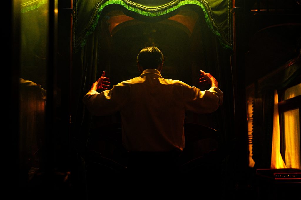 A person stands with their back to the camera in a dim, golden-lit room, arms raised and slightly outstretched as if addressing or conducting an unseen audience. They wear a white shirt and are framed by heavy green curtains, which create a dramatic, almost theatrical atmosphere. The lighting casts deep shadows, giving the scene an intense and mysterious mood.