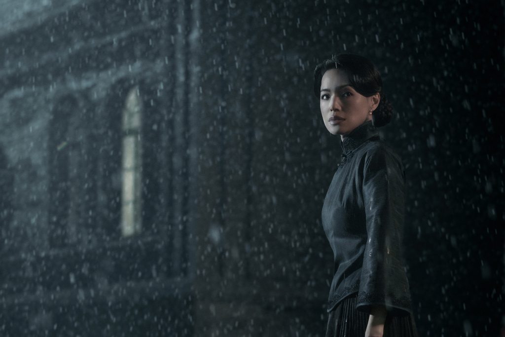 A person wearing a dark traditional-style outfit stands outdoors at night in falling rain, looking toward the camera with a calm but serious expression. Their hair is neatly styled, and an old stone building is blurred in the background, giving the scene a dramatic, atmospheric mood.