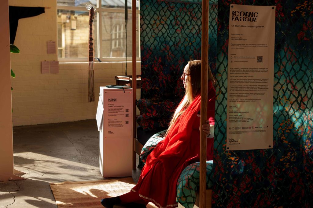 Woman in a red dress sits on a bench covered in material with a green scale pattern on. She looks at the the window where the sun comes through casting shadows into the room. 