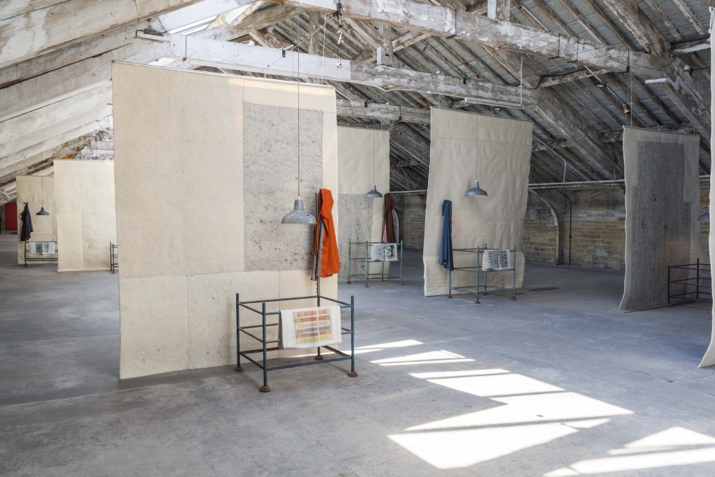 A large, airy industrial loft with exposed wooden beams and a slanted roof, where tall fabric panels hang from the ceiling like soft partitions. Metal lamps hang above minimal metal stands holding fabric samples, with a few garments draped on the panels. Sunlight streams across the concrete floor, creating bright rectangular patches in the open, gallery-like space.