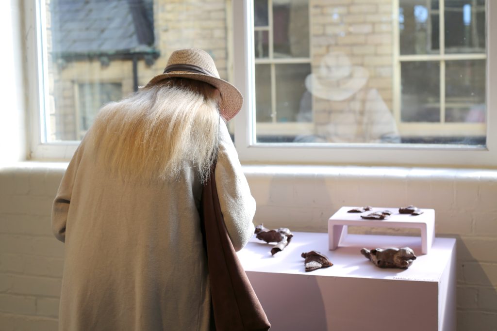 A visitor in a long coat and hat stands over a table covered by a baby pink cloth. On the table is a variety of chocolate horse models of different size and shape. 