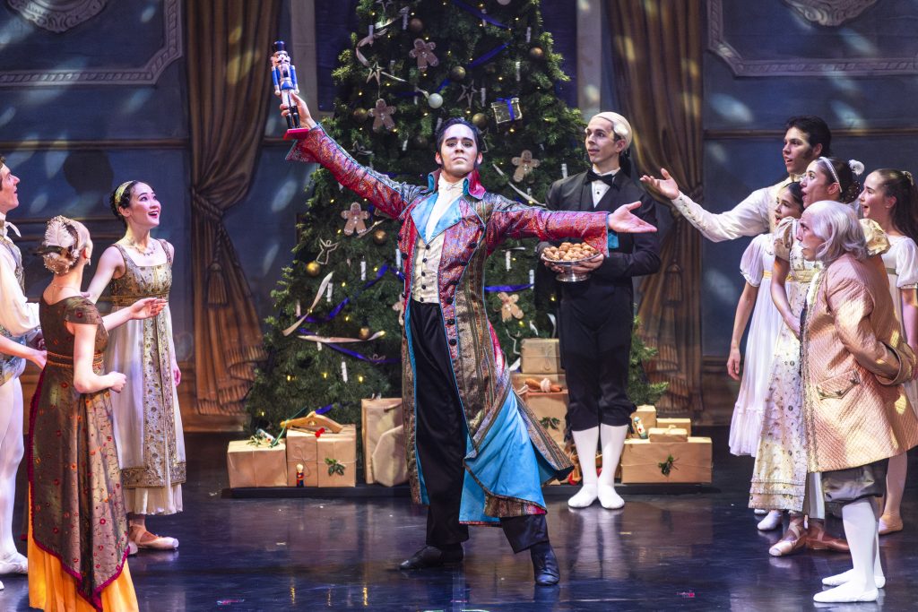 A performer portraying Uncle Drosselmeyer stands centre stage holding up a nutcracker doll, surrounded by dancers in period costumes gathered around a decorated Christmas tree, capturing a lively Christmas party scene from The Nutcracker.