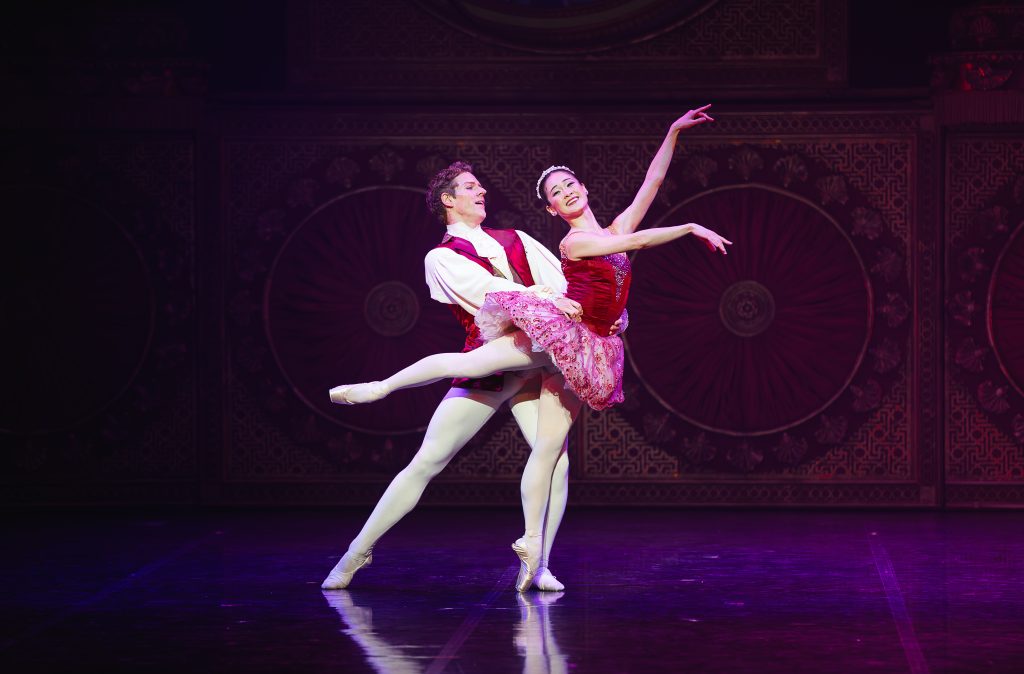 The Sugar Plum Fairy and her Cavalier perform an elegant partnered lift on stage, dressed in rich pink and red costumes, captured mid-dance against a dark, ornate backdrop during The Nutcracker.