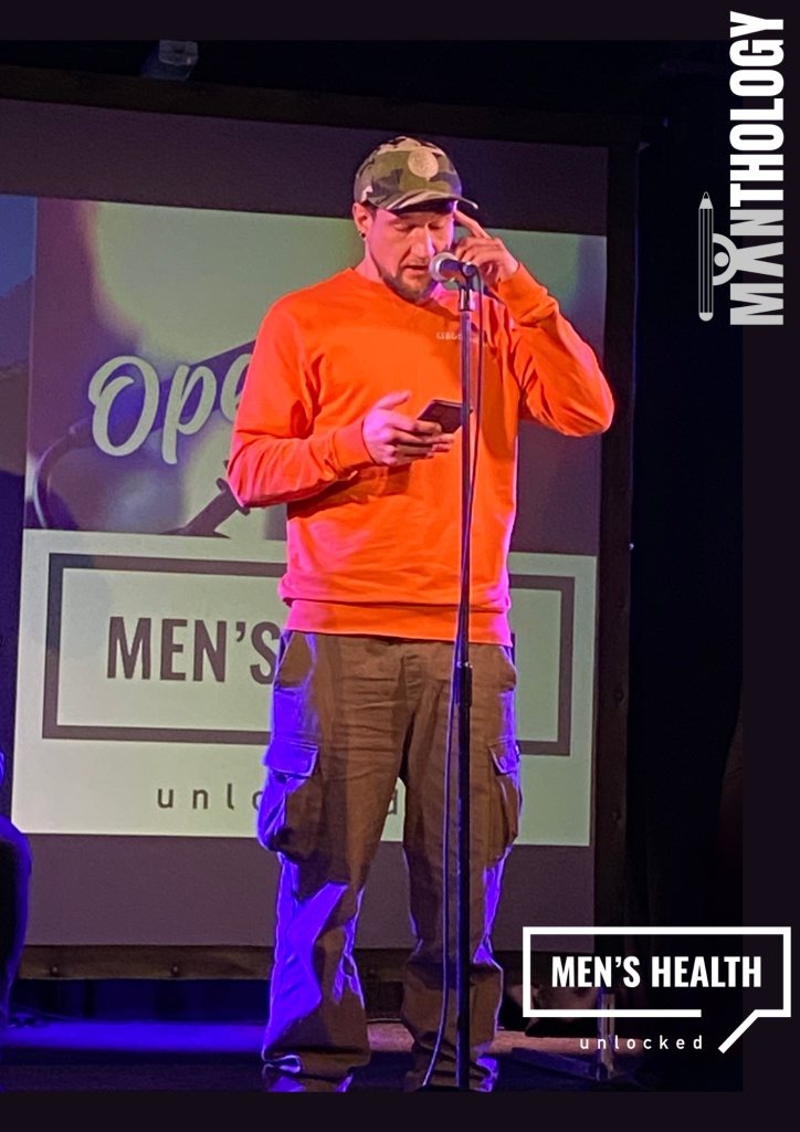 A man in an orange top stands at a microphone, reading from his phone on a dimly lit stage, with “Men’s Health Unlocked” displayed behind him.