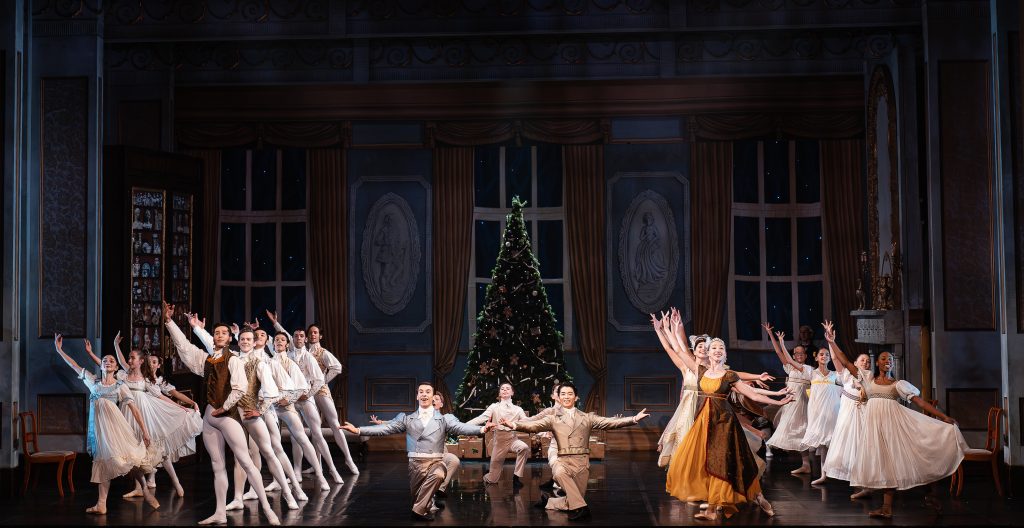 Ballet dancers perform a joyful Christmas party scene on stage, arranged in elegant poses around a decorated Christmas tree inside an ornate grand living room, with performers in period costumes celebrating in unison during The Nutcracker.