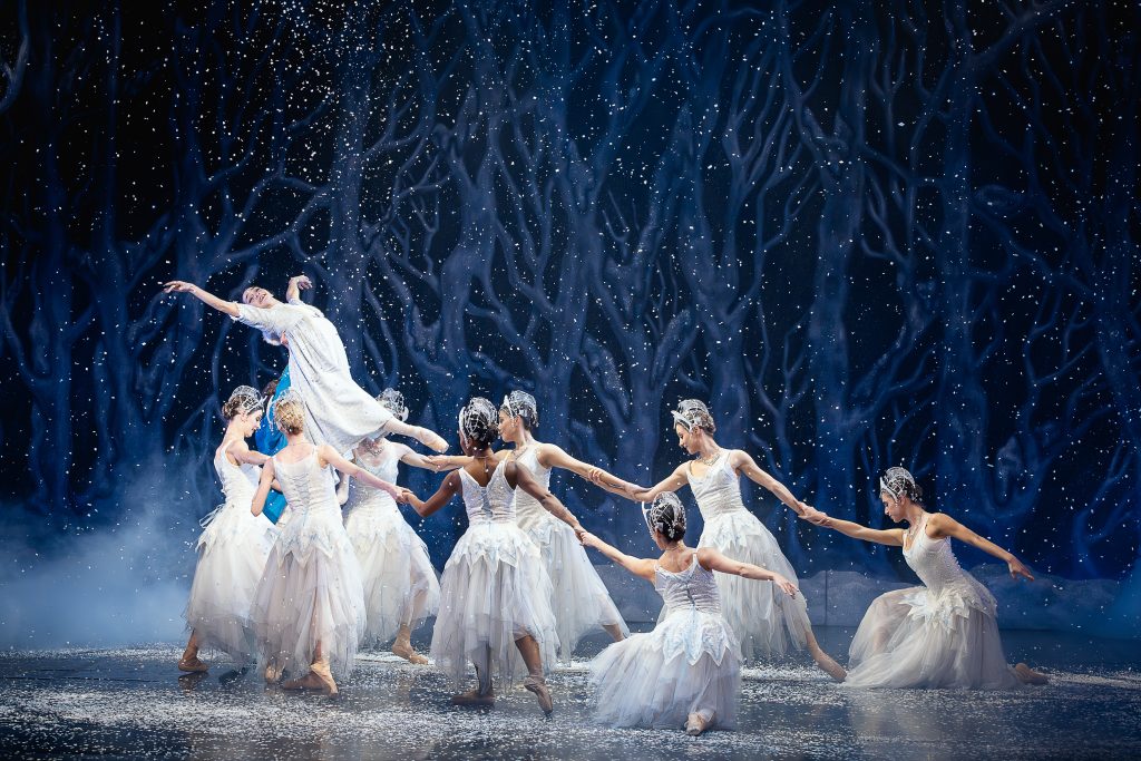 Ballet dancers in white, snow-inspired costumes perform a graceful ensemble lift beneath falling snow, set against a dark blue winter forest backdrop, evoking a magical snowy woodland scene from The Nutcracker.