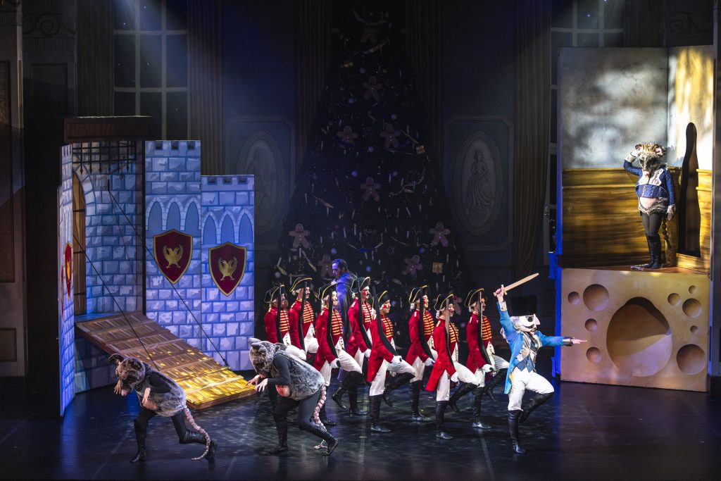 Ballet dancers perform a dramatic battle scene featuring toy soldiers in red uniforms, mischievous mice in grey costumes, and the Mouse King on a raised platform, set against an oversized Christmas tree and castle backdrop in The Nutcracker.