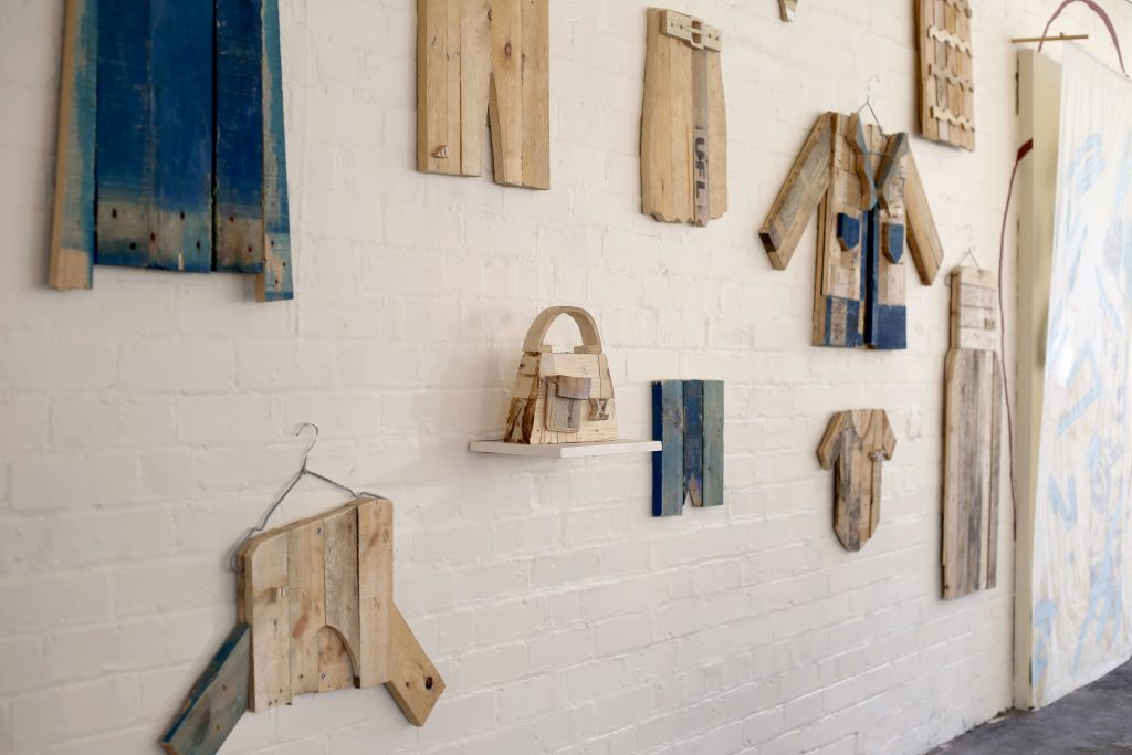 Image of brick wall painted white. On the wall are hung wooden pallets designed to look like designer clothes, from a jacket to a handbag. 