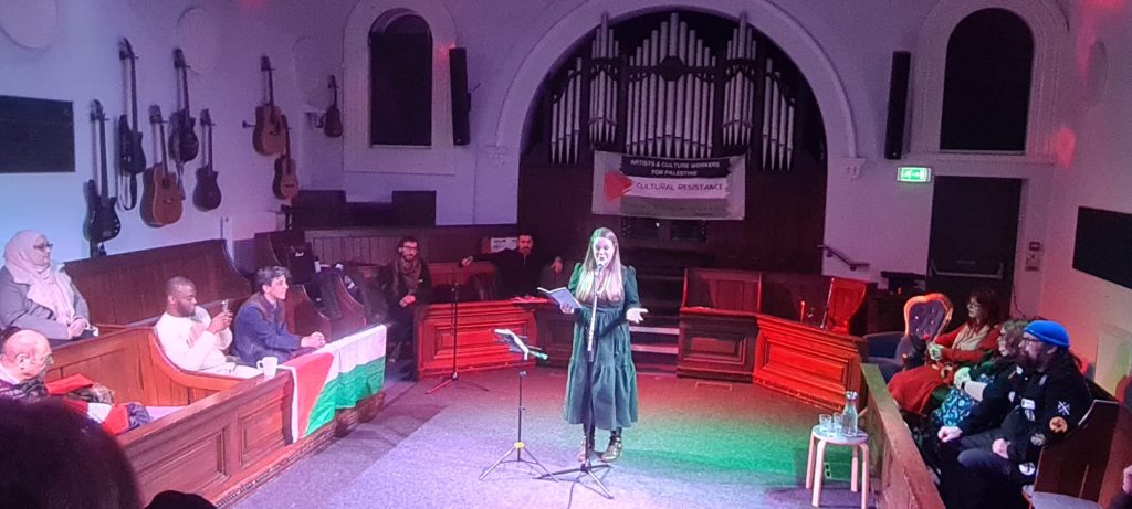 A woman reads from a page at a microphone in a church-like hall, with an audience seated around her and guitars hanging on the walls.