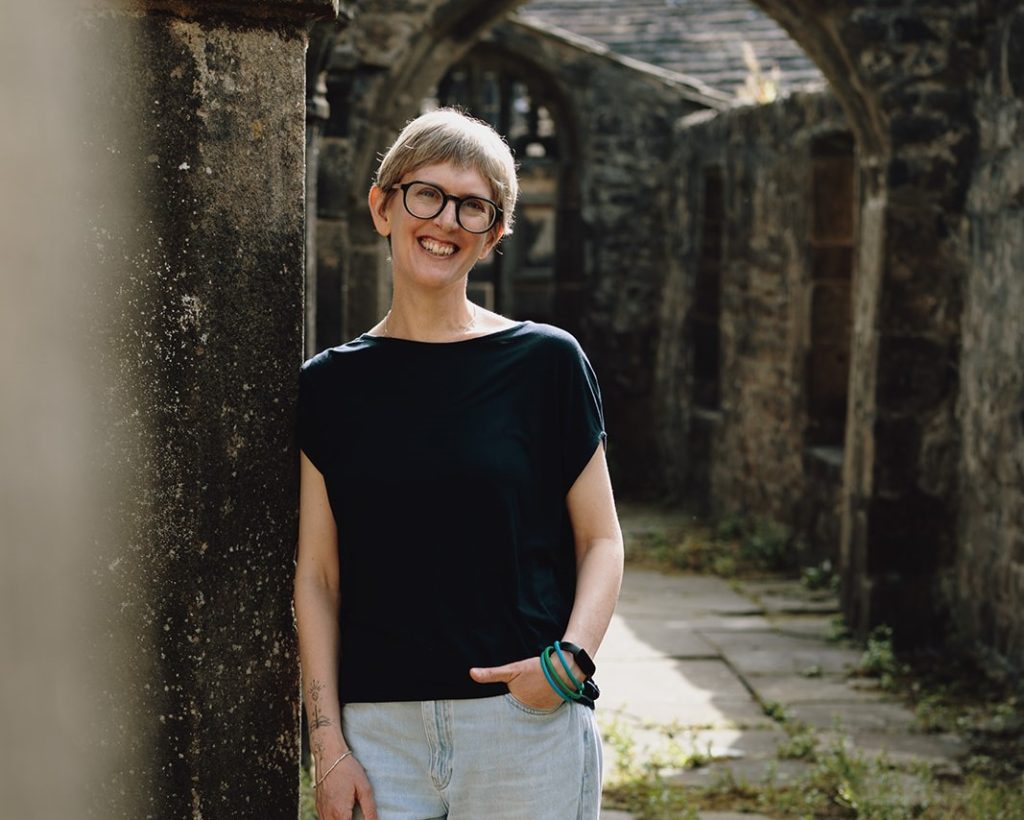 A smiling person with short grey hair and glasses stands in a stone courtyard, wearing a black top and light blue jeans, with historic stone buildings and archways in soft focus behind them.