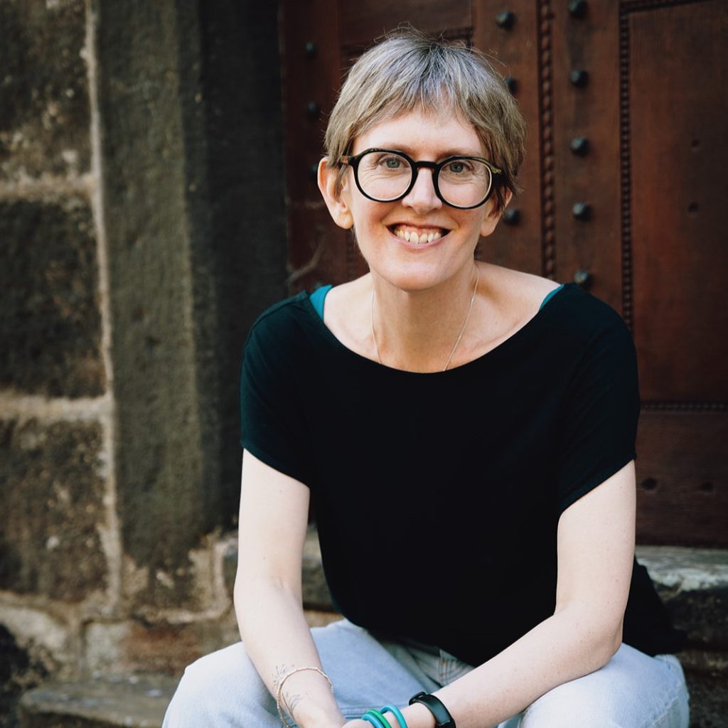 A smiling person with short grey hair and glasses sits on stone steps in front of a large wooden door, wearing a black top and light blue jeans.