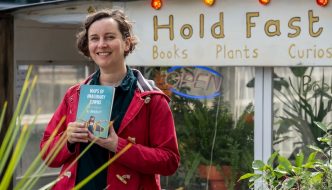 SJ Bradley stands outdoors in front of a small shop called “Holdfast” holding a book titled Maps of Imaginary Towns. They are wearing a red jacket and standing among potted plants. A neon “Open” sign glows in the shop window behind them, and warm string lights hang along the roofline.