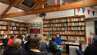 Audience seated in a cozy bookstore with wooden beams and shelves of books, listening to a woman speaking at the front beside a table of books.