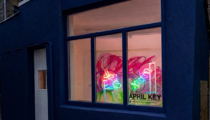 Small, dark-blue shopfront at dusk with a large window displaying colourful neon light sculptures in pink, green, and blue. The glowing installation sits against a painted backdrop inside the compact gallery space, with “APRIL KEY” visible on the window.