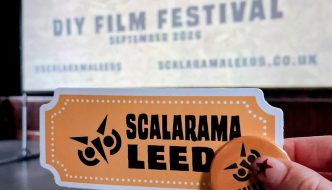 Hand holding a Scalarama Leeds sticker and badge in front of a projected screen displaying “Scalarama Leeds DIY Film Festival – September 2026”.
