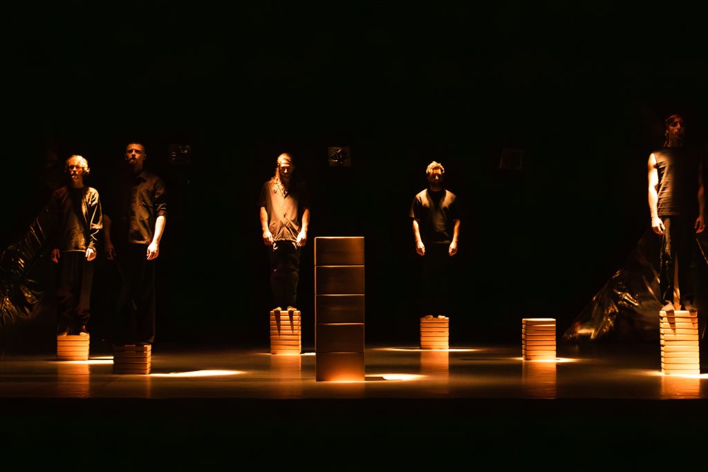 Five performers from 'Tomorrow' cast stand spaced apart on stacked blocks under warm spotlights on a dark stage.