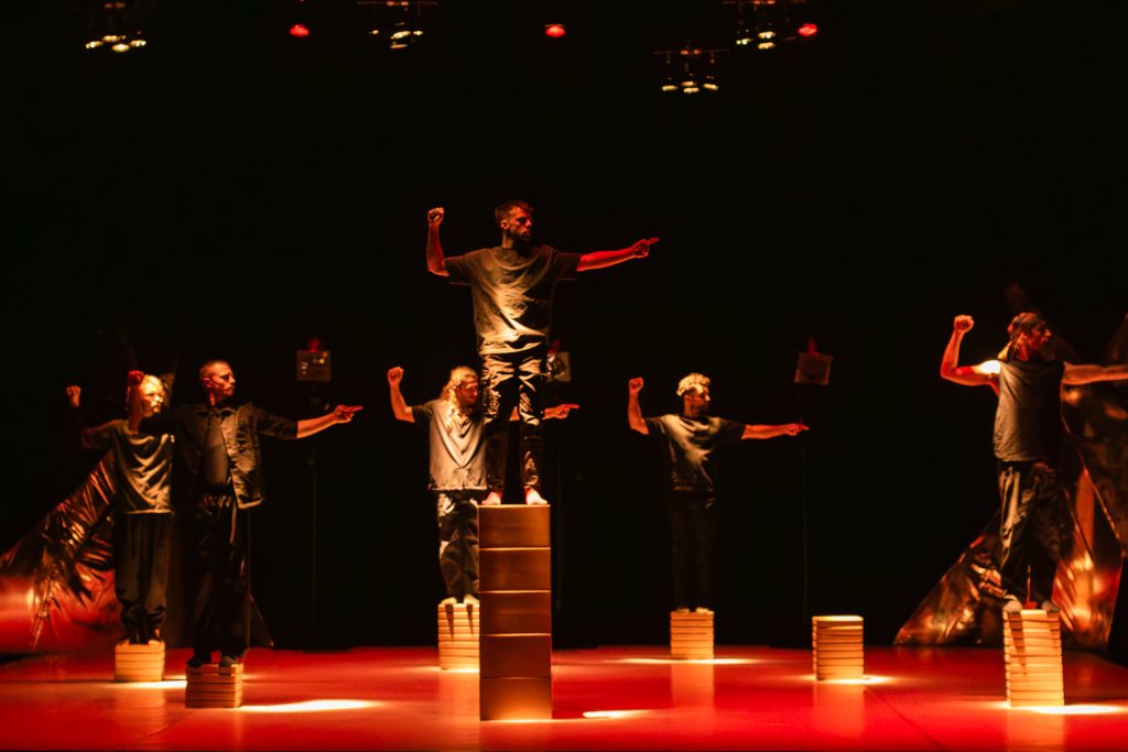 Dancers from the 'Tomorrow' cast pose on stacked platforms under dramatic red stage lighting in a contemporary performance.