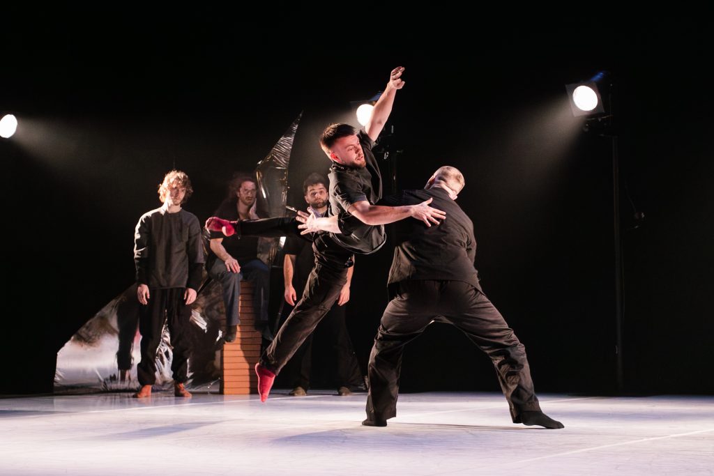 A dancer lifts another mid-air on a dim stage, with three performers watching in the background under bright lights.