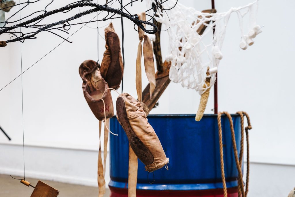 Worn ballet shoes hang from tangled netting and ropes above a blue oil drum, alongside knotted cords and suspended found objects in a gallery installation.