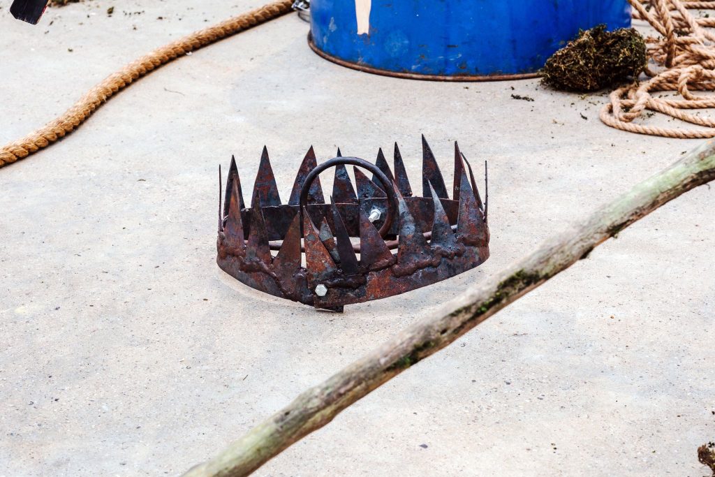 A rusted bear trap sits open on the gallery floor like a jagged crown, surrounded by rope, driftwood and debris near a blue oil drum.