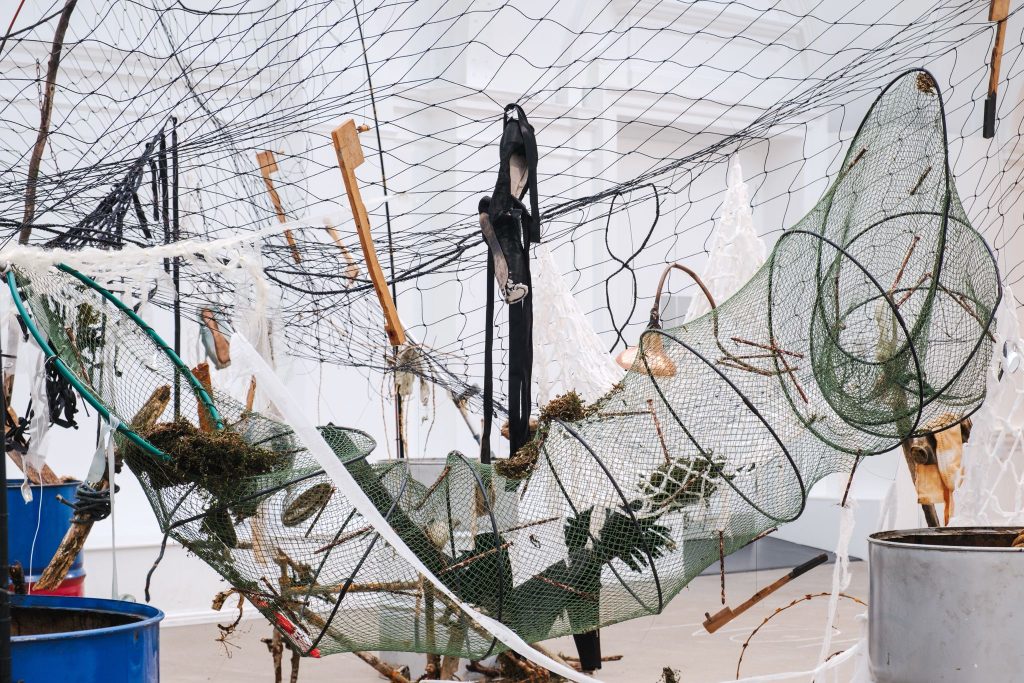 Green dredging nets sag between rods, filled with sticks, debris and organic matter, suspended within a web of black and white netting in the gallery space.
