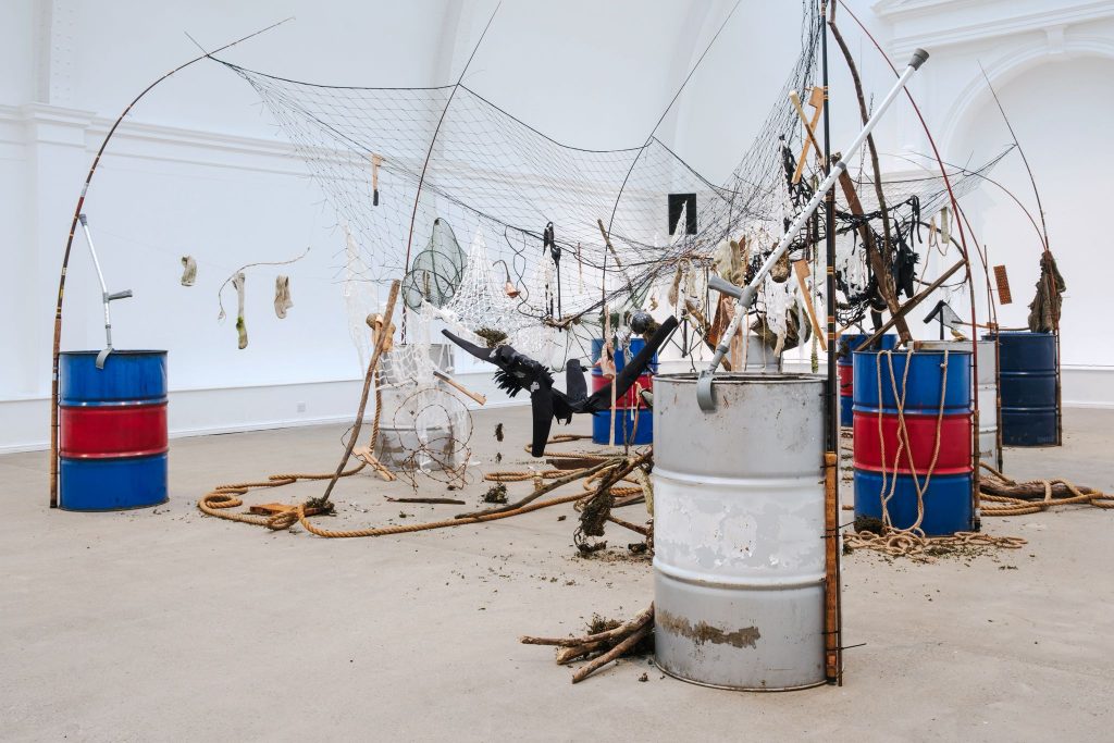 A sculptural arrangement of oil drums, fishing rods and suspended nets filled with found objects—ropes, clothing, driftwood and debris—forming a fragmented canal-boat shape in a gallery space.