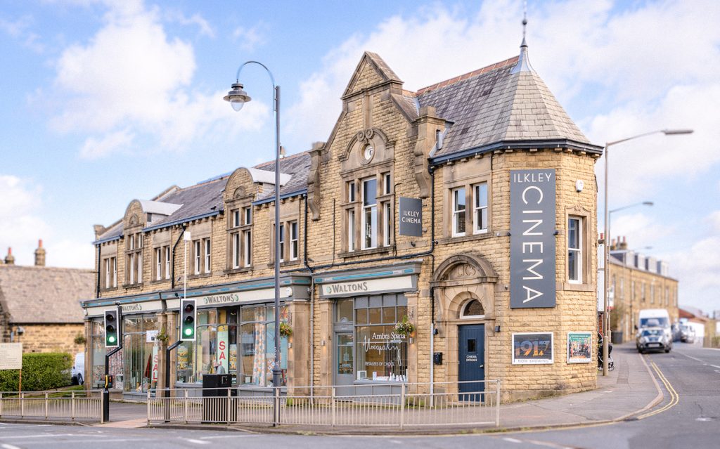 Exterior of Ilkley Cinema, a stone building on a street corner with large windows, shopfronts at ground level, and a tall vertical “Ilkley Cinema” sign on the façade.