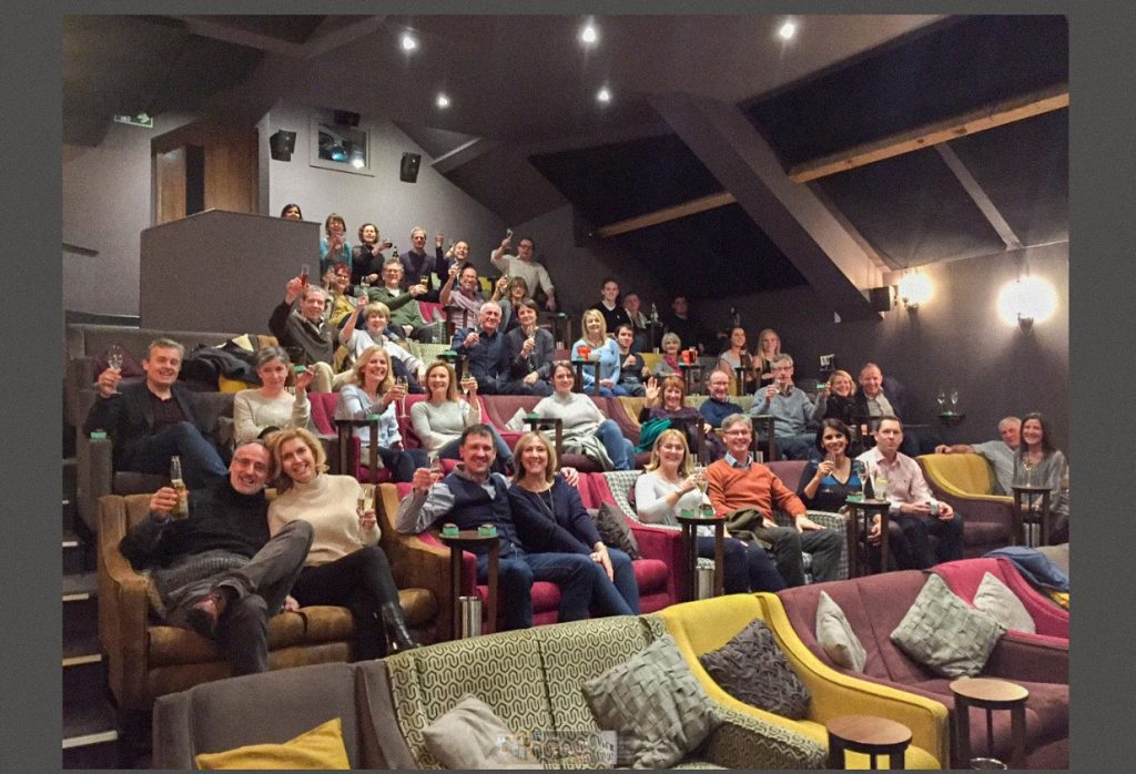 Audience seated in a boutique cinema with tiered sofas and armchairs, smiling and raising drinks toward the camera in a relaxed, social atmosphere.