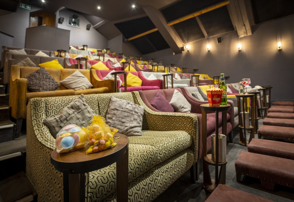 A cozy boutique cinema with tiered seating featuring plush sofas and armchairs in warm tones, decorated with colorful cushions. Small side tables hold snacks like popcorn, drinks, and candy, creating an intimate and comfortable movie-watching atmosphere.